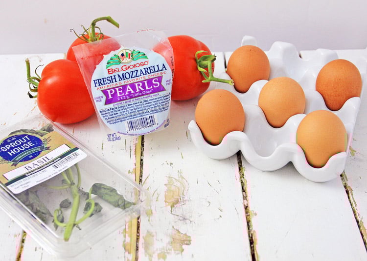 Ingredients for a caprese quiche laid out on a white table top including eggs, mozzarella pearls, tomatoes, and basil.