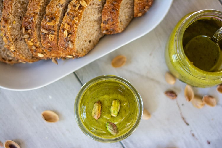 top down view of pistachio butter in a jar with a loaf of sliced bread and shells sprinkled on the table.