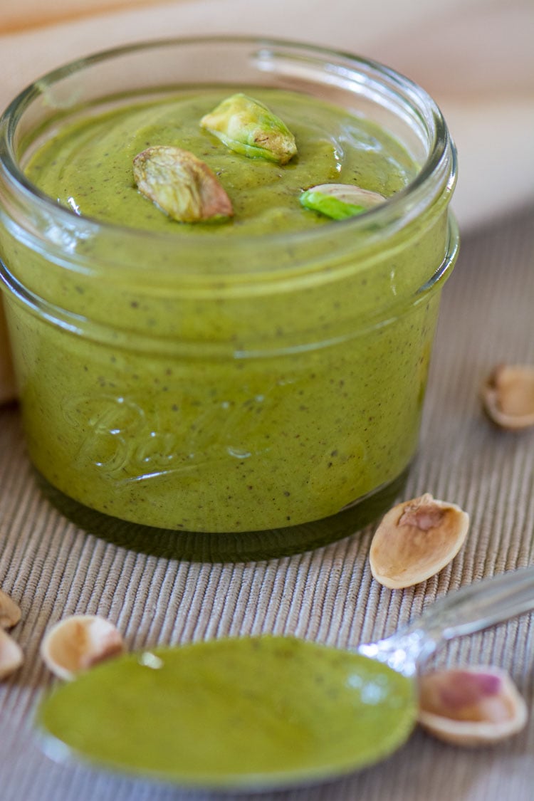 homemade pistachio butter in a jar with a spoon and shells on the grey tablecloth.