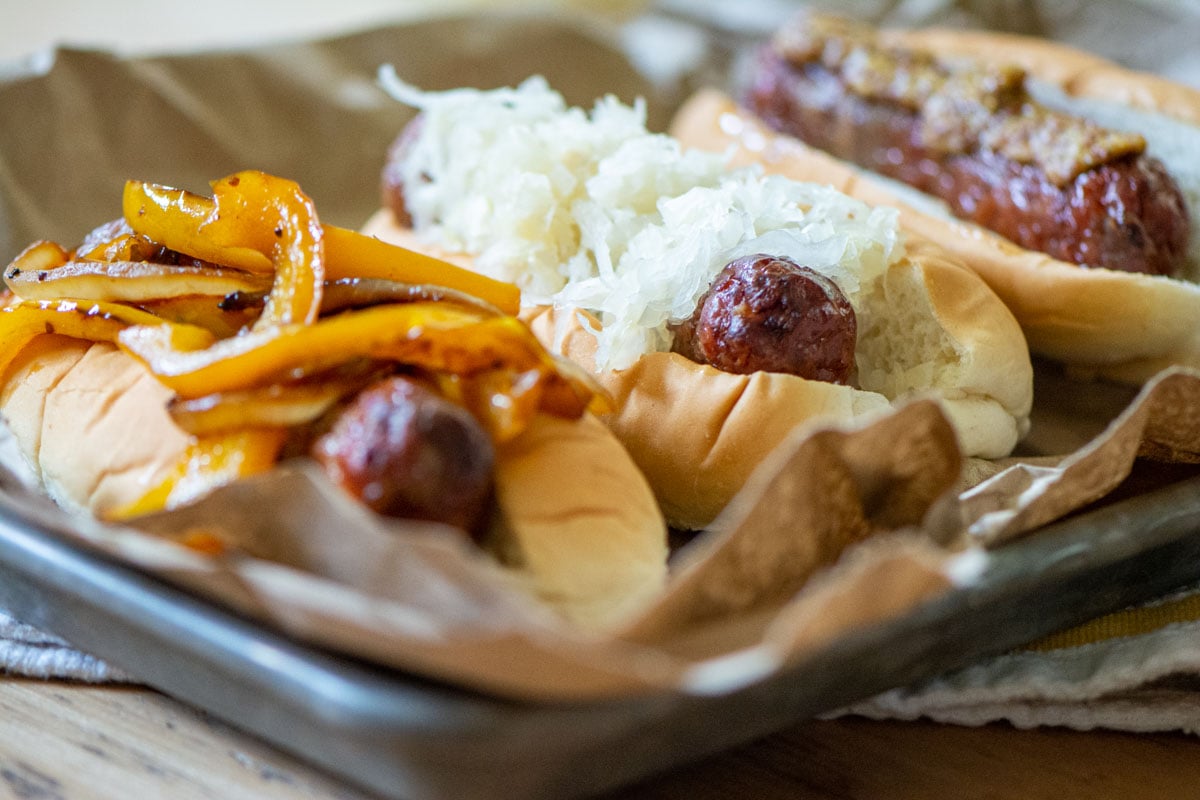 three brats on a small sheet pan. One is topped with onions and peppers, one has sauerkraut, and one has whole grain mustard.