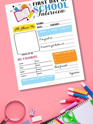 Free printable first day of school interview on pink surface next to colorful school supplies.