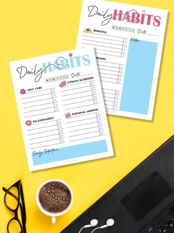 Free printable daily habit trackers on a yellow surface next to coffee cup and reading glasses.