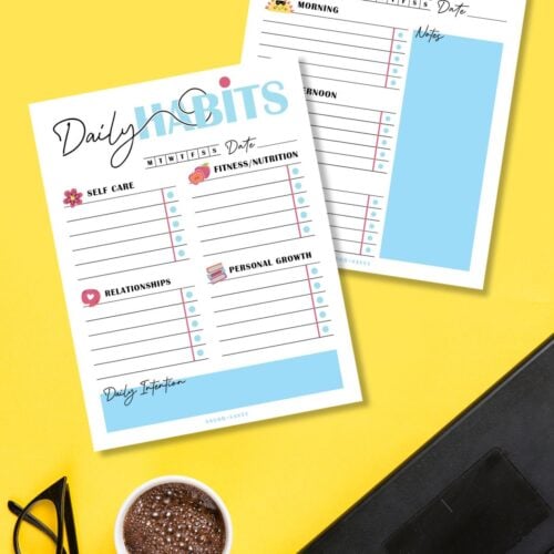 Free printable daily habit trackers on a yellow surface next to coffee cup and reading glasses.